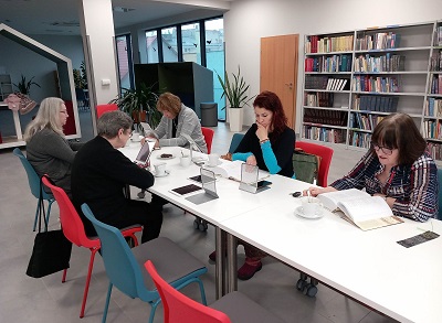 5 kobiet siedzi przy stole w czytelni, czyta korzystając z książek i tabletów